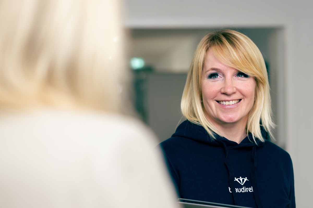 Auf dem Bild sind zwei blonde Frauen zu sehen. Die eine sieht man nur von hinten, Die andere lächelt und trägt einen blaudirekt Hoodie,