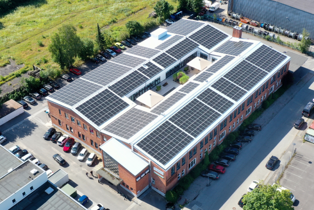 Ein riesiges Gebäude mit Solaranlagen auf den Dächern, zugeparkte Autos drumherum, und einer Grünfläche links.