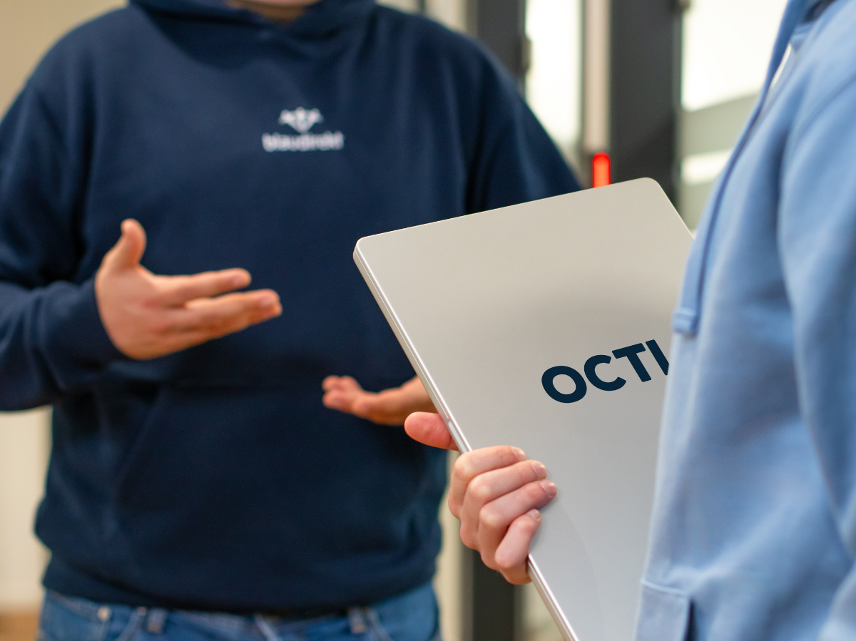 2 Menschen stehen in einem Gespräch gegenüber. Einer hält einen Lapotop mit der Aufschrift "OCTI" in der Hand.