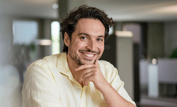 Ein Mann sitzt an einem Tisch, hält die Hand am Kinn und lächelt in die Kamera.