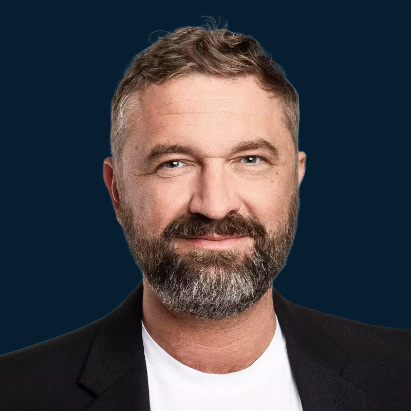 Ein lächelnder Mann mit Bart, hinter ihm ist ein blauer Hintergrund.