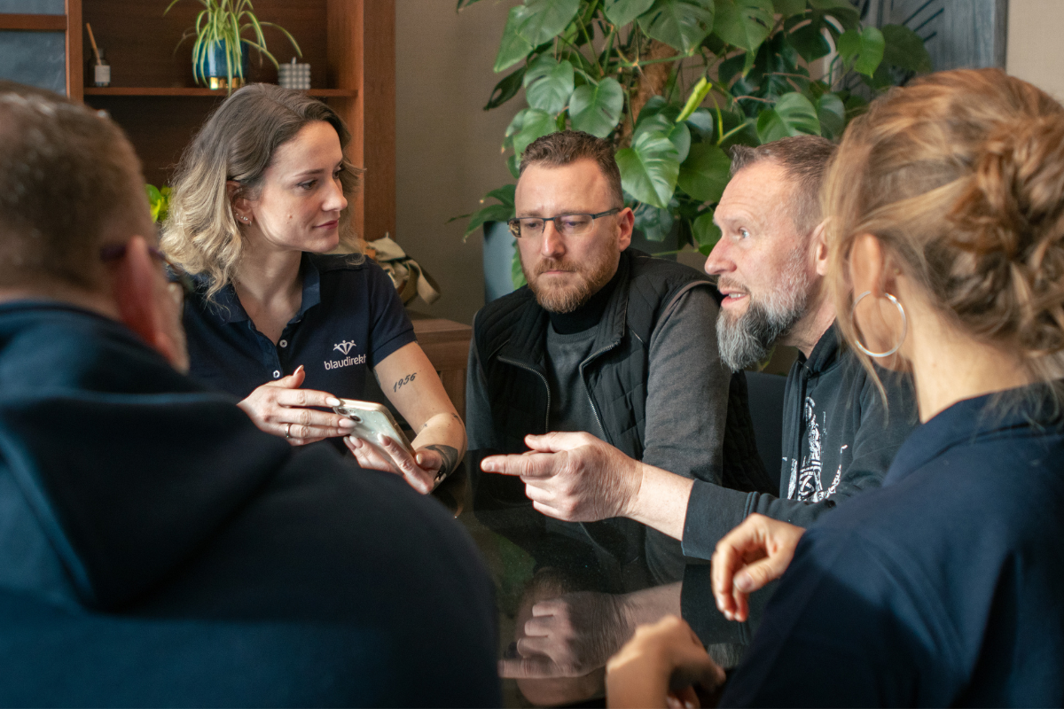Es sind fünf Mitarbeiter in einer Besprechung zu sehen. Sie befinden sich im Office und man sieht Pflanzen hinter ihnen.