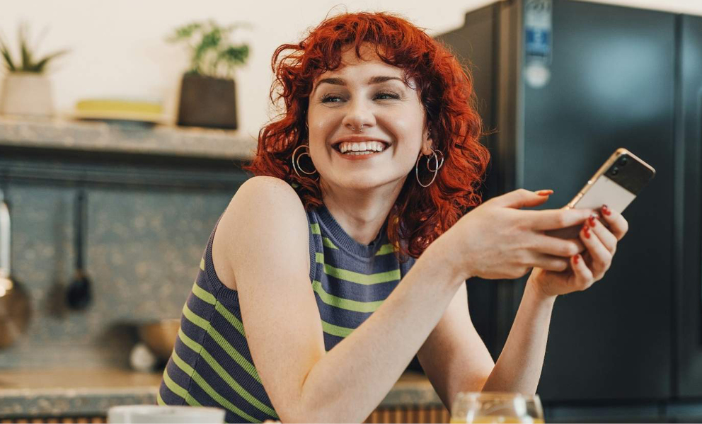 Eine lächelnde rothaarige Frau mit einem Handy auf den Händen. Sie befindet sich in einer Küche.