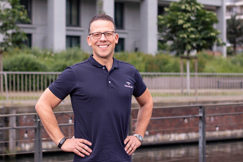 Im Vordergrund ist ein lächelnder Mann mit einer aufrichtigen Haltung zu sehen. Er trägt ein blau direkt Polo T-Shirt. Im Hintergrund sieht man ein Gebäude und Bäume.